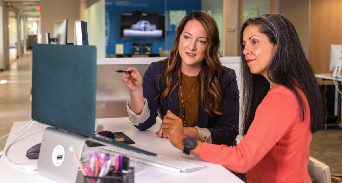 Two business women talking about sales in office at desk with laptop