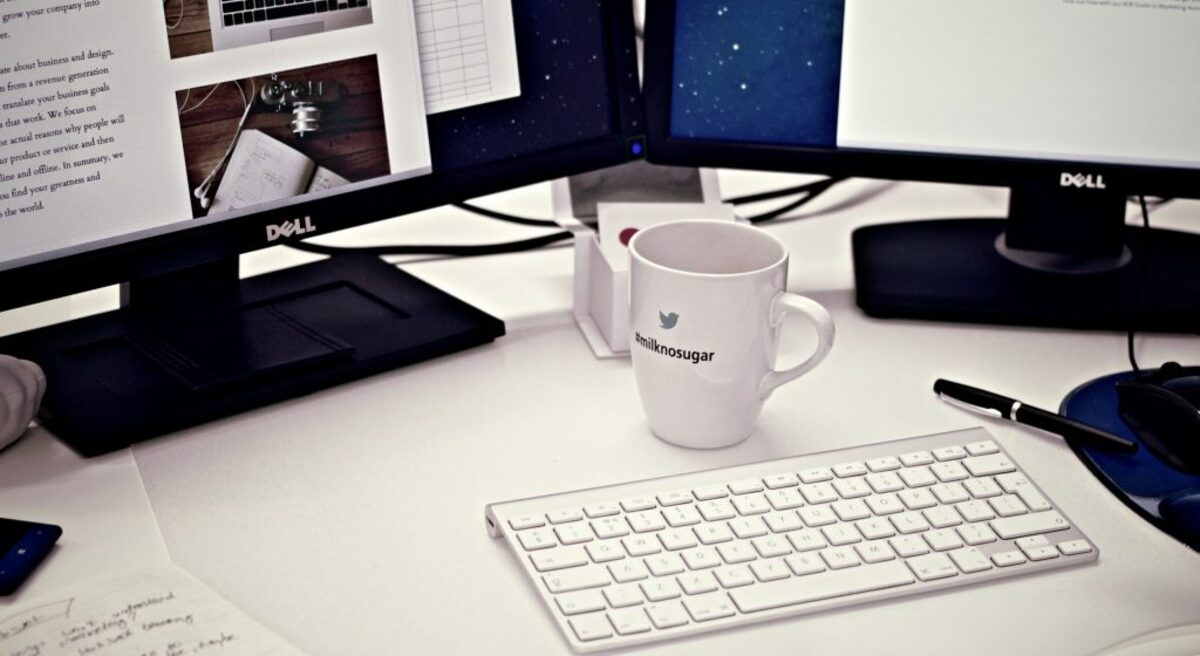 White ceramic mug between apple magic keyboard and two flat screen computer monitors