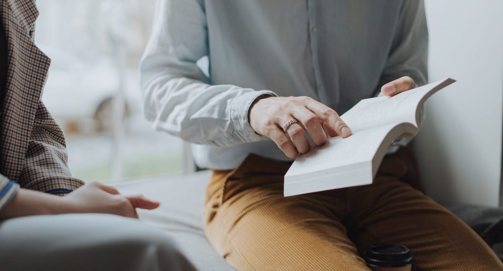 Man in white dress shirt and brown pants sitting on white chair reading book