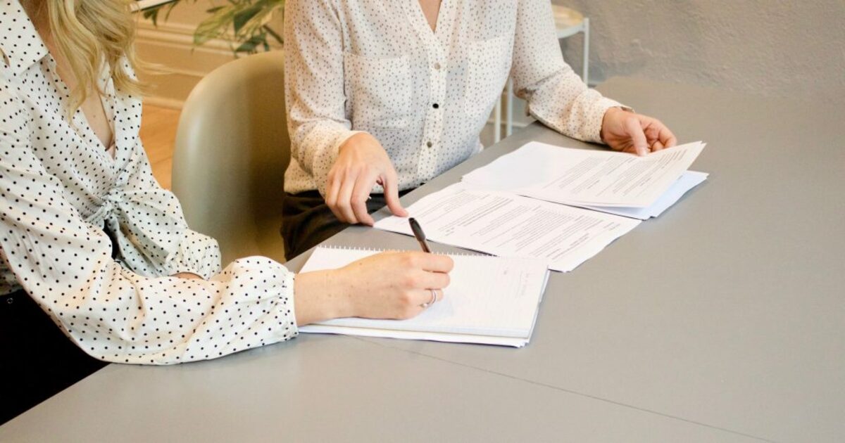 woman signing on white printer paper beside woman about to touch the documents