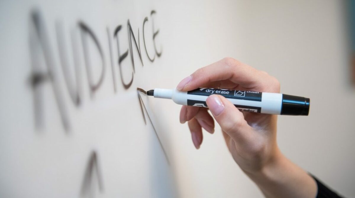 Woman's hand writing the word "audience" on a whiteboard, with arrows.