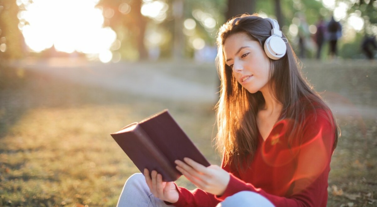 Selective focus photo of smiling woman in a red long sleeve top reading book while listening to music on headphones